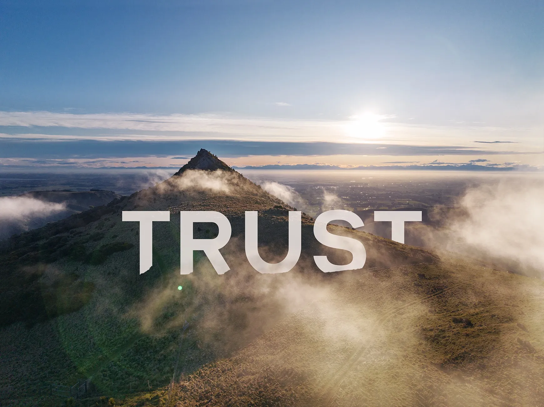 Gibraltar rock, Christchurch NZ- the top peaks up above white fluffy clouds and there is a stunning sunset in the distance - the word trust written in white peaks out, visible just below the peak
